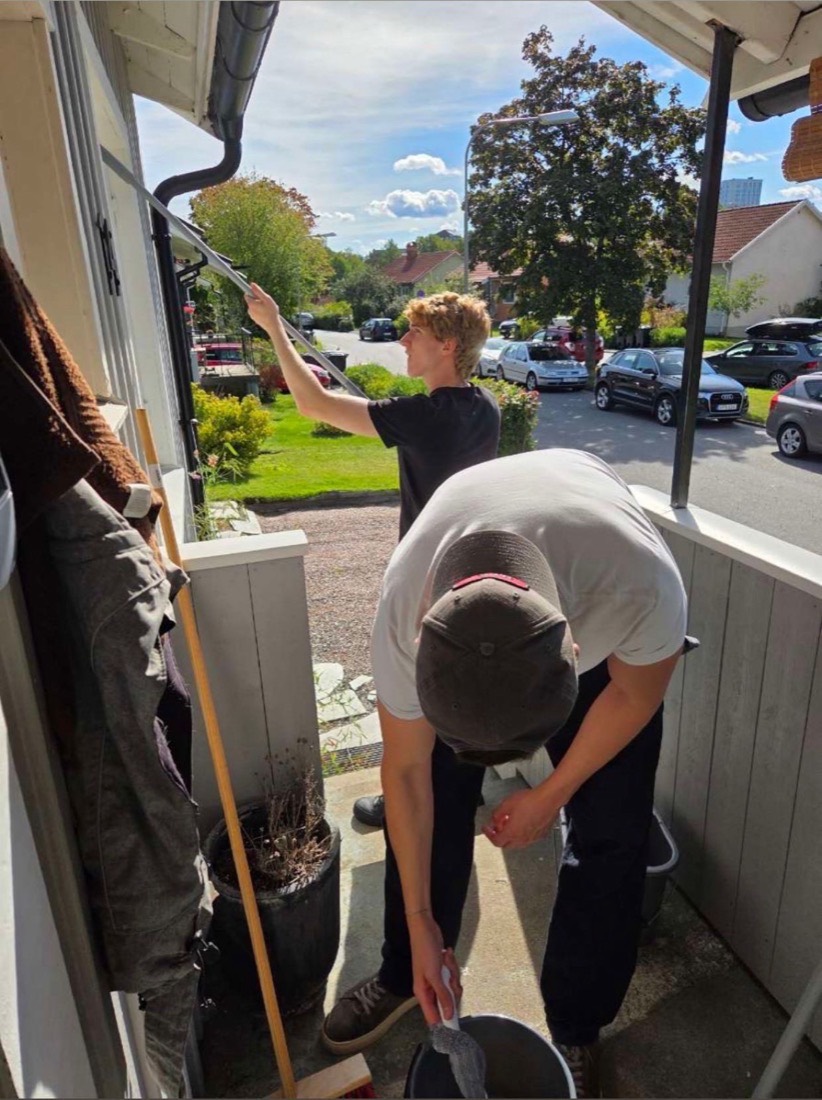 Två av oss putsar fönstren på en villaveranda i Stockholm, en med skrapa på stång och en som vrider ur duken